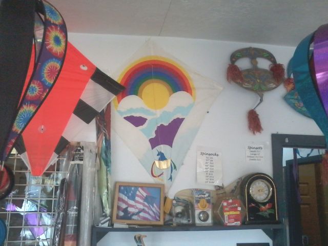 A picture of the cluttered inside wall of a kite store, the last Catch the Wind in Pacific City on the Oregon Coast. In the center of the photo, a little hard to see since it is a partially white kite on a white wall. It is a unicorn; riding in the golden basket of a hot air balloon; with a print of a rainbow, the sun, and clouds on the balloon, on a kite. The golden basket has 4 colored streamers from it, with swallow tails.