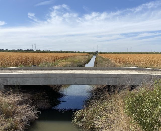 A concrete bridge spans a narrow waterway surrounded by golden fields and scattered vegetation under a blue sky with wispy clouds. Electric power lines are visible in the background.