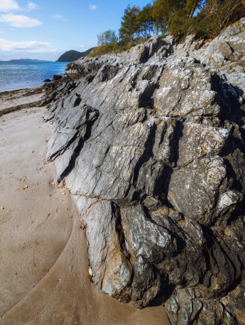 The rugged beauty of Benderloch beach at Ardmucknish Bay in Argyll & Bute, Scotland. The foreground is dominated by a dramatic outcrop of weathered, grey rock formations, their layers and fissures etched by time and tide. These rocks contrast sharply with the smooth, golden sand of the beach. In the background, the calm blue waters of the bay stretch towards distant, tree-covered headlands under a partly cloudy sky. The scene exemplifies the raw, untamed nature of Scotland's coastline, where ancient rocks meet the sea in a timeless tableau of natural beauty.
