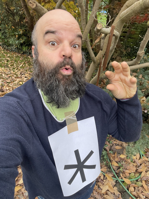A person with a beard and a bald head is making a funny surprised expression outdoors, wearing a dark sweater. They have a large white paper with a black symbol taped to their shirt. The background features foliage and trees with fallen leaves.