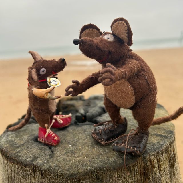 Photo of Minimus and Silvius, the Latin mice, standing on an old breakwater with the North Sea behind them. Minimus is trying to shake Silvius' paw, but Silvius is playing a trick on him by wearing a crab claw that he found among the seaweed and shells