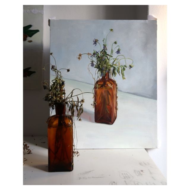In the foreground is an orange glasses bottle with dried flowers inside. Behind and oil painting with the same motif.