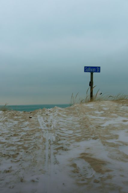 We're back in January of this year. There's fresh snow on the ground. We're at the northern terminus of Sullivan St, in Gary Indiana, where the road meets the sand and then the sand meets Lake Michigan. We're looking up a path over the dune, where tan-and-white snow/sand covers the bottom third of the photo, and uniformly milky, overcast skies cover the top chunk, and in between, at the horizon, a sliver of emerald colored water shows over the top of the dune. 