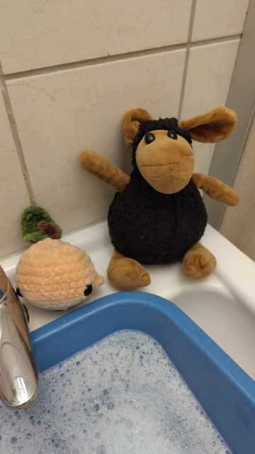 Ein schwarzes Plüschschaf und ein Pfirsichwal, die am Rand eines Waschbeckens sitzen. Im Waschbecken sieht man eine blaue quadratische Schale, in der Wasser mit Waschmittel eingelassen ist.