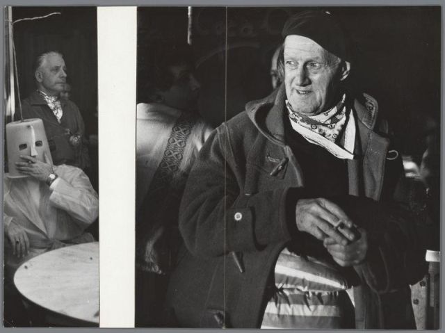 The image is a black and white photograph depicting two elderly men in what appears to be an indoor setting with dim lighting. The man on the right side of the photo, closer to the camera, has visible wrinkles on his face and wears glasses; he's dressed warmly in layers: a coat over another garment featuring intricate embroidery or lace at its neck area. Around his neck is a patterned scarf, possibly indicating colder weather attire. His headgear appears to be a beret, adding to an air of formality.

He holds what seems like a walking stick firmly with both hands and has a contemplative expression on his face as he looks off towards the left side where another man sits in partial view behind him; this individual is wearing a mask that covers half their head. The environment suggests they may be at some kind of public event, hinted by other indistinct figures visible in the background.

The photograph has been digitally manipulated or altered since it shows two nearly identical men on either side with slight variations - one appears more faded than the others which could indicate age-related discoloration when printed but is not indicative of digital enhancement. The overall composition and setting suggest a historical context, potentially documenting cultural events from an earlier era, as indicated by clothing styles and grayscale photography typical for older publicatio [...]