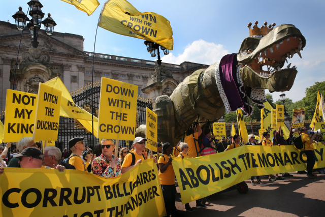 A vibrant anti-monarchy protest taking place in front of Buckingham Palace under a clear blue sky. A large group of demonstrators, many wearing yellow shirts, hold bright yellow placards and banners with slogans like "NOT MY KING," "DOWN WITH THE CROWN," and "ABOLISH THE MONARCHY." The focal point is a massive, satirical puppet of a Tyrannosaurus Rex wearing a gold crown and a royal purple-and-ermine mantle, symbolizing the monarchy as a "dinosaur." In the foreground, a man in a colorful patterned shirt looks on, while protesters hold a large yellow banner that reads "CORRUPT BILLIONAIRES..."

