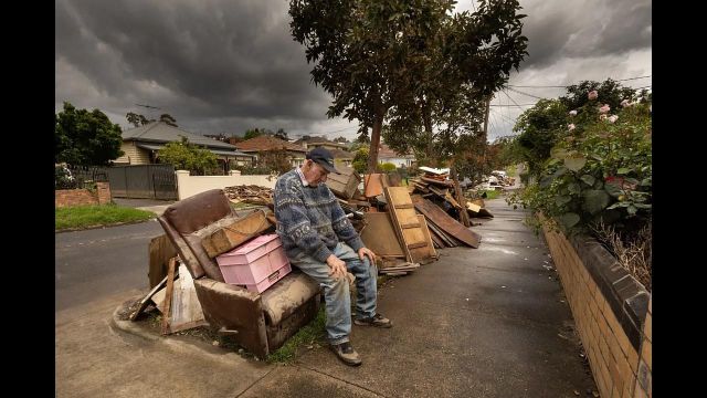         86-Year-Old Brian Matthews (Jason South)
        
An elderly man sits limply on a ruined armchair on the curb, his head down, chin tucked to his chest, surrounded by the debris of his flooded home under a bruised sky. This posture represents the final stage of chronic psychological strain: the total physical and nervous collapse of despair once a threat has struck and destroyed everything.