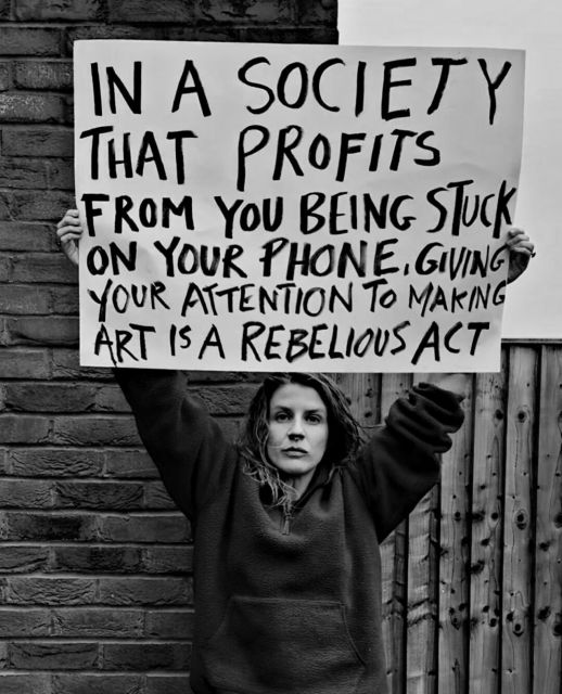 A woman holding a sign saying: In a society that profits from you being stuck on your phone, giving your attention to making art is a rebellious act.