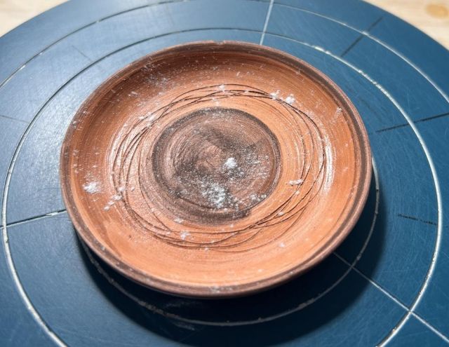 A small round orange plate on a blue banding wheel, with darker iron oxide stains in circles, and white powder (borax) sprinkled on top.