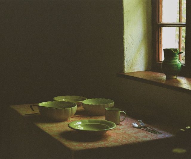 A table covered in a reddish patterned cloth stands in a dark room, lit only by a small window in the upper right corner. On the cloth sit three pale green scalloped bowls, a green enamel mug and a shallow green plate at the front, with two forks lying together at the right and the handle of a ladle resting in the left-hand bowl. The window has a brown-red frame and a thick, roughly plastered reveal that catches soft daylight; a green glazed jug with a chipped rim stands on its deep sill. Beyond the reveal the wall falls into near-black shadow, and the light on the tablecloth fades to dark toward the left edge. Colour photograph on 35mm film, with visible grain.